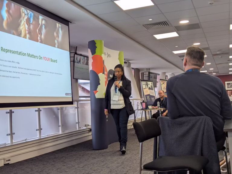A presentation slide on a large screen reads 'representation matters on your board'. A woman in a suit is speaking into a microphone near the screen, several men are visible sitting around tables.